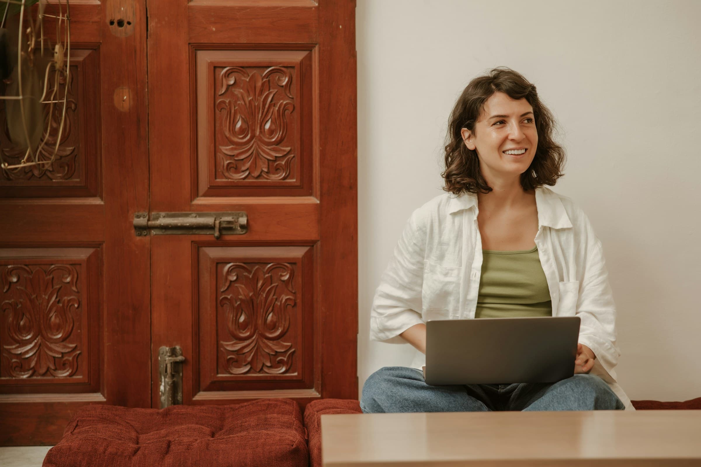 A woman sits with her laptop on her knees, smiling, beside a carved wooden door