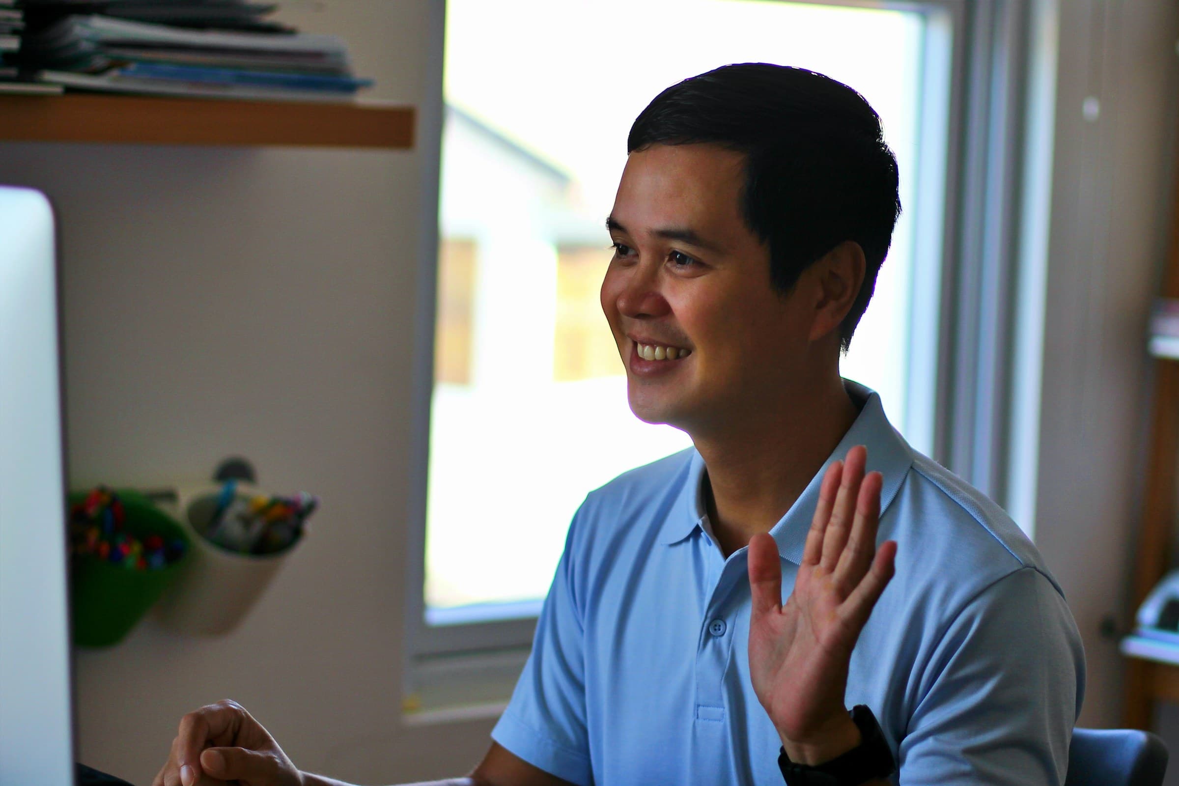 A man at his desk smiles and waves at his screen during a video call