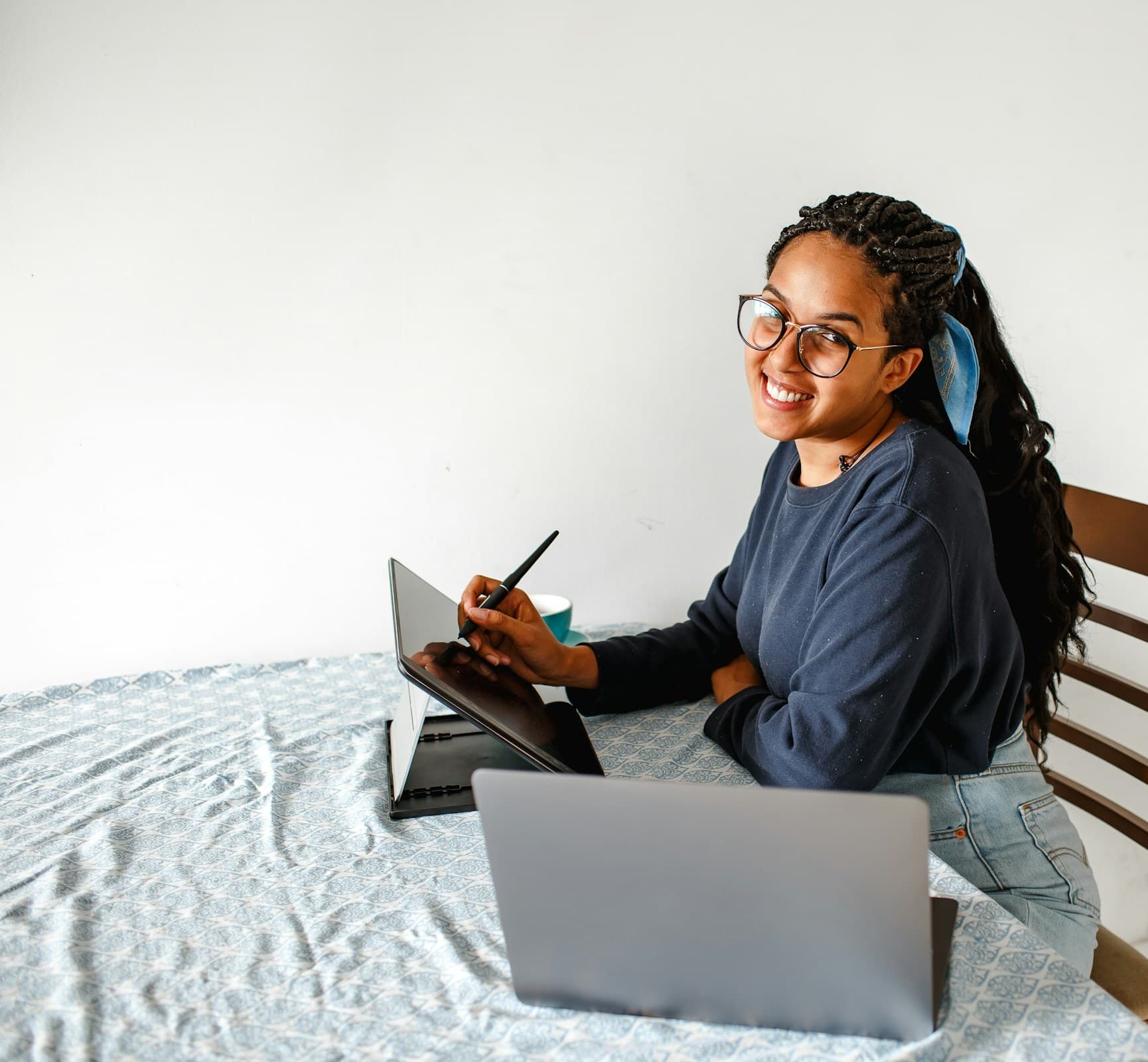 A woman smiles up from her laptop and tablet at a table at home