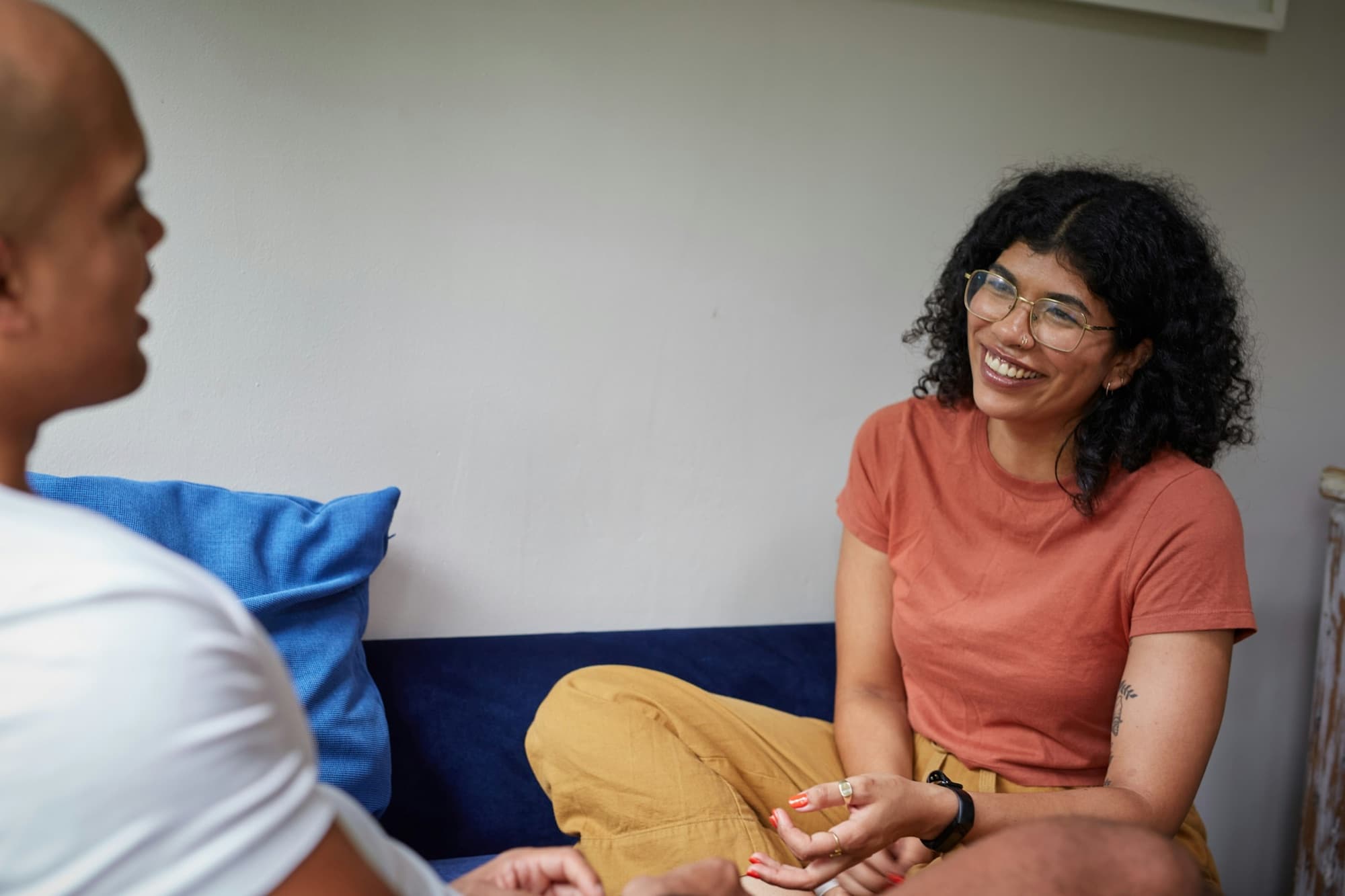 A woman in glasses laughs mid-conversation with a man on a blue couch